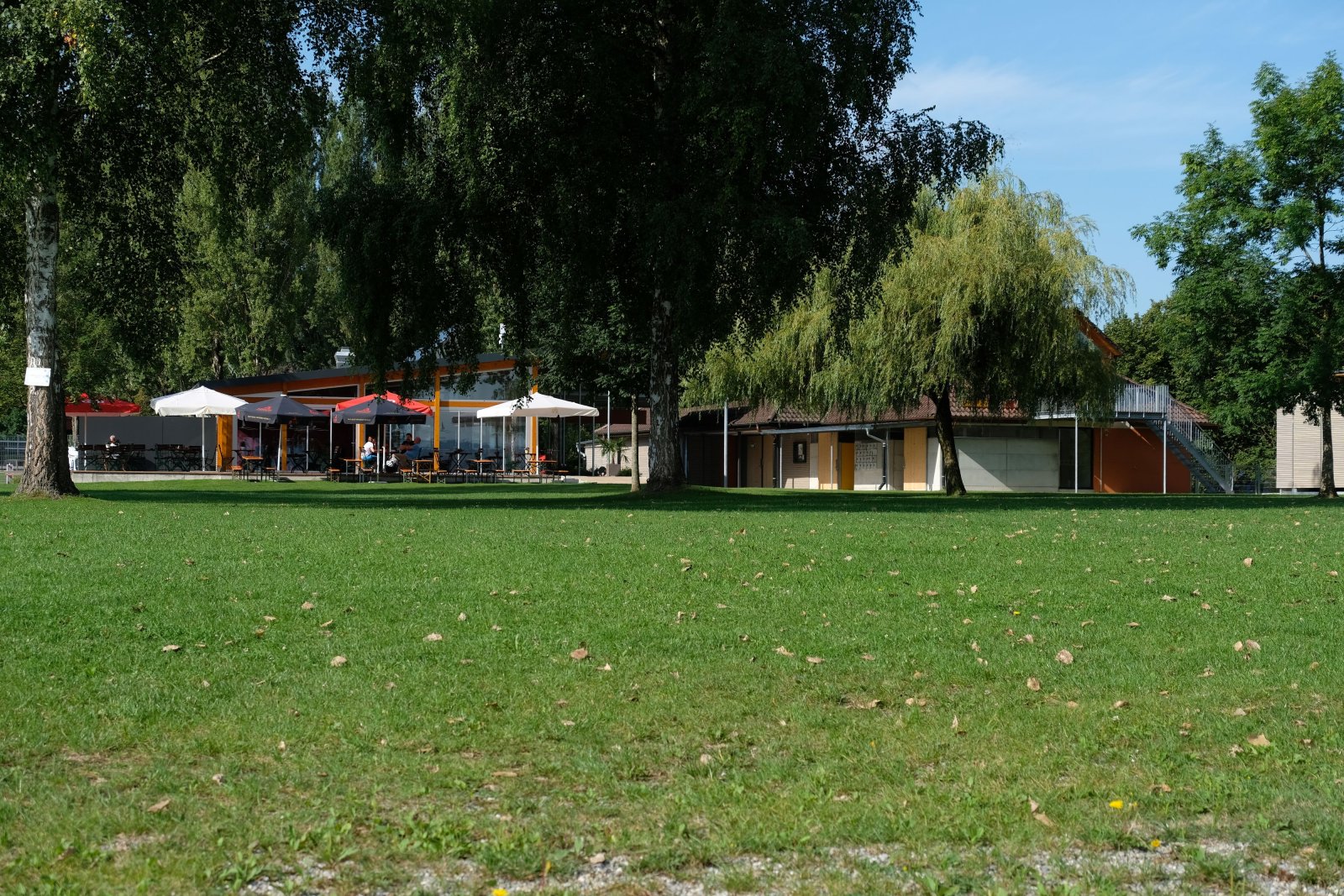 Tatsächliches Strandbad Iznang: Liegewiese mit Schattenbäumen, Strandbadgebäude und Außenterrasse.