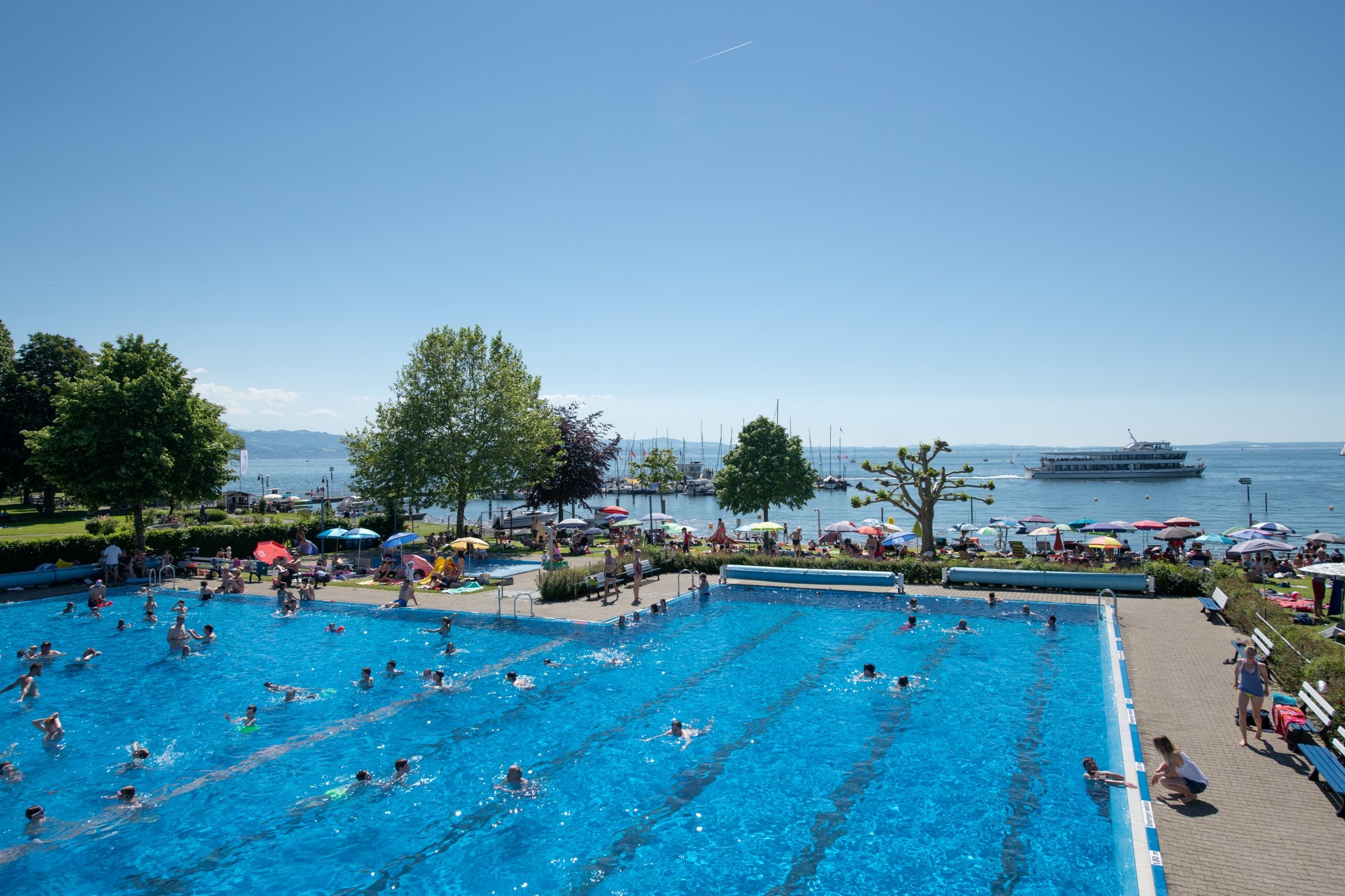 Großes Freischwimmbecken des Strandbads Nonnenhorn mit Badegästen und Bodensee im Hintergrund